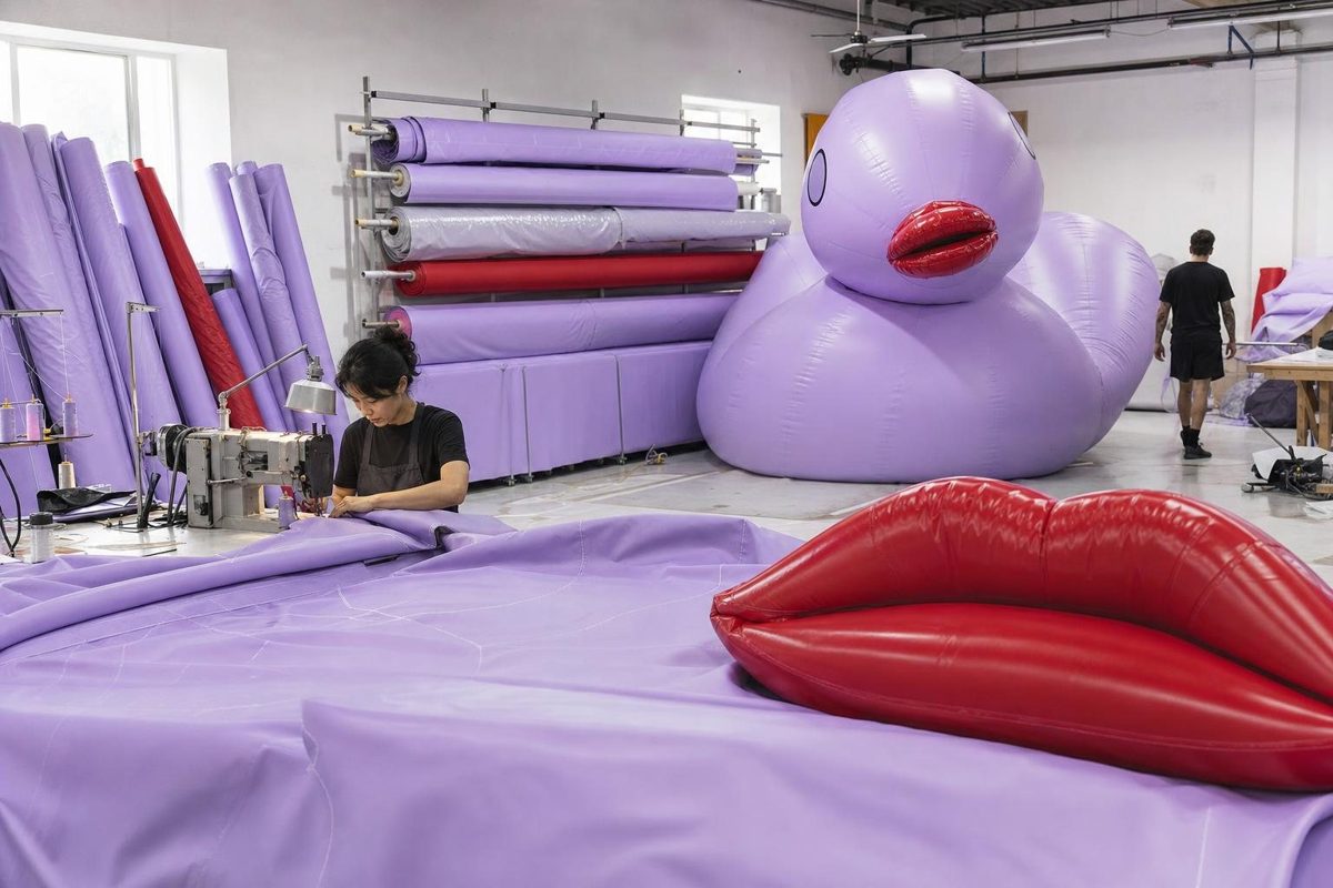 Lavender fabric panels being sewn in an inflatable workshop, with the red lips piece and the duck body behind